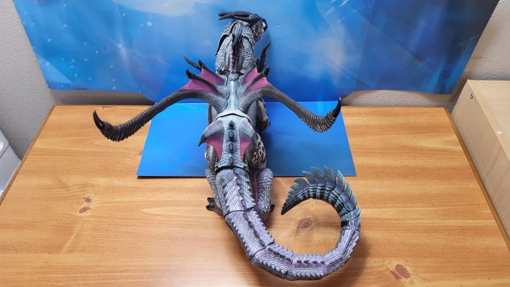 A look at the figure from the backside, and also above. Tail is curled on the right as much as possible. Jaw on the figure is closed, and the head and neck is slightly facing to the right. Figure is in front of the included package backdrop.