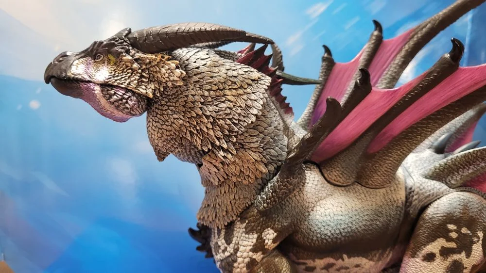 A close up of the left side view of the figure with head, neck, wings, and torso in view. Figure is in front of the included package backdrop. Jaw is closed.