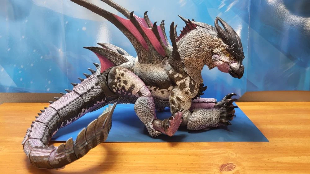 Figure is in a sitting posture. Right side view. Neck is posed down, and slightly to the right. Jaw is closed. Tail is curled on the right as much as possible. Figure is in front of the included package backdrop.