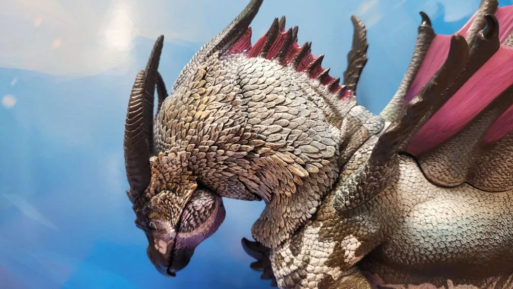 A close up of the figure's head and neck from the left side view. Figure is in front of the included package backdrop. Jaw is closed. Neck is positioned as low as possible.