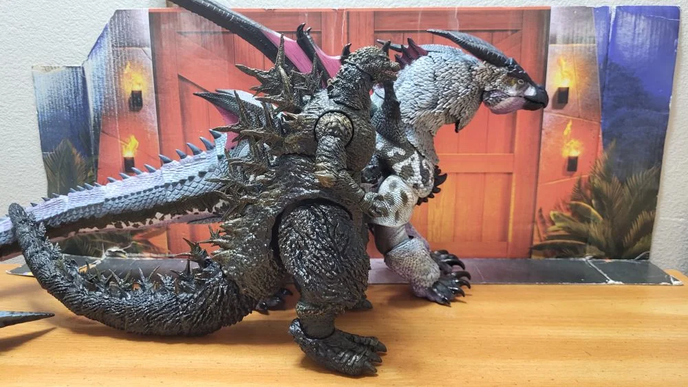 Comparison with the Super7 Ultimates Minus One Godzilla. Both figures from a right side view. The Godzilla is in front of Kuraokami.. Both in a neutral pose with jaws closed. In front of the reviewer's typical JW background.