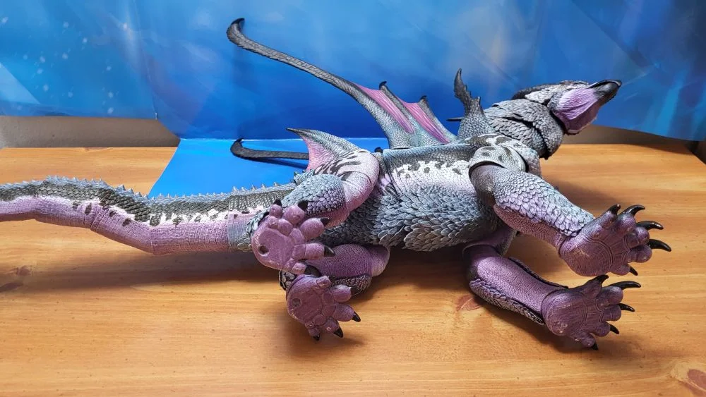 A look at the underside of the figure, mostly the paws, belly, and some of the tail. Figure is laid down on its left side, with the head facing the right. Jaw is closed. Figure is in front of the included package backdrop.